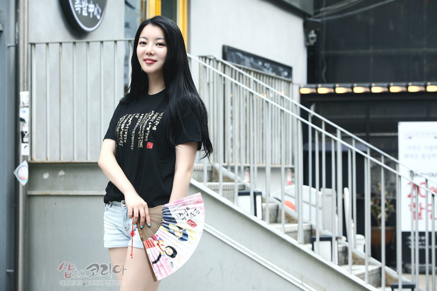 [텍스트: 샵오브코리아] 검은색 면 티셔츠 위 금색 입체 발포 기법으로 훈민정음 서체와 붉은 낙관 문양을 세로 배열한 디자인