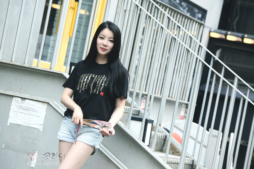 [텍스트: 샵오브코리아 대한민국의 숨겨진 가치를 전합니다] 검은색 면 티셔츠에 금색 입체발포 기법으로 표현된 세로 배열 훈민정음 서체와 붉은색 사각 인장 포인트가 조화된 디자인 이미지 설명