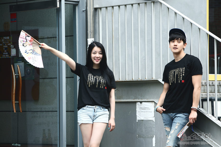 [텍스트: 훈민정음 한글 서체 문구] 검은색 면 티셔츠 위에 금색 입체 발포 기법으로 새긴 훈민정음 서체가 세로 배열된 캐주얼 반소매 티셔츠 연출