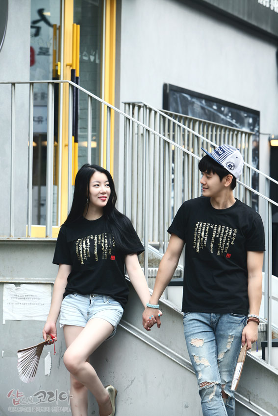 [텍스트: 샵오브코리아 대한민국 전통문화 전자기기 전자기기] 흑색 면 티셔츠 위에 금색 입체 발포 기법으로 표현된 세로 배열 훈민정음 한글 서체와 붉은색 전각 도장 포인트가 돋보이는 커플 연출 이미지