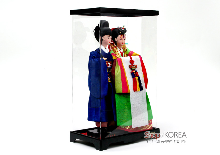 [텍스트: Shop of KOREA, 대한민국의 품격까지 전합니다.] 비단 한복 입은 신랑신부 인형을 투명 케이스에 담은 장식, 색동 원삼과 자수 노리개 매듭, 선명한 청색 도포와 녹색 치마가 어우러진 전통 혼례 인형 작품