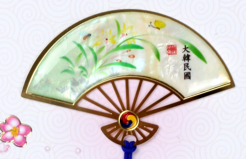 [텍스트: 大韓民國] 금속 테두리에 자개 상감된 반달 부채형 책갈피, 옻칠 바탕 위 난초와 나비 문양, 태극 문양 장식과 청색 끈 매듭 구성