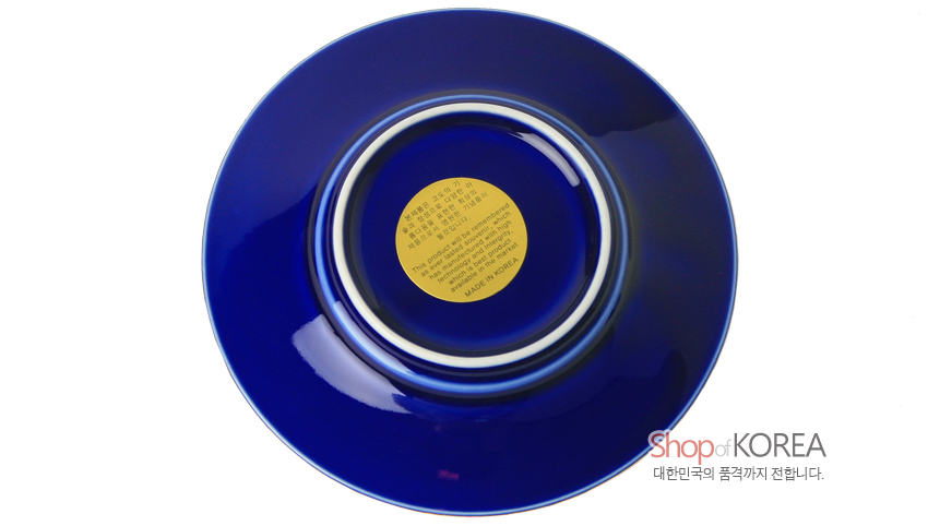 [텍스트: Shop of KOREA 대한민국의 품격까지 전합니다. MADE IN KOREA] 짙은 청색 바탕 위 중앙에 금색 원형 라벨이 부착된 금속 재질 장식 접시 뒷면 모습