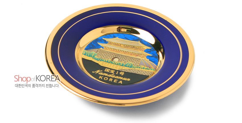 [텍스트: Shop of KOREA / 대한민국의 품격까지 전합니다. / 國寶1号 Namdaemun KOREA] 청색 바탕 위 금속 재질에 24K 금장으로 남대문 전경을 입체 양각 표현하고, 가장자리에 이중 금색 띠 장식을 더한 원형 장식 접시 디자인