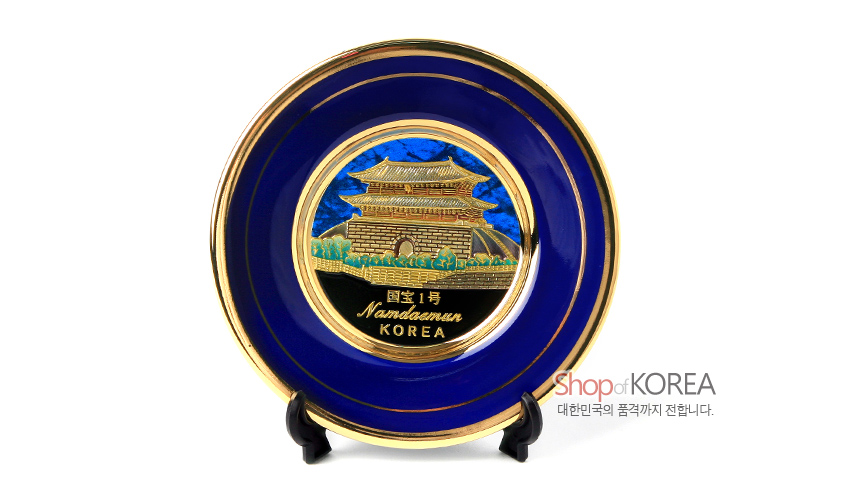 [텍스트: 國寶1號 Namdaemun KOREA] 청색 바탕 위 금속 재질에 24K 금장 남대문 전경을 양각으로 표현하고 금색 이중 테두리를 두른 원형 장식 접시 모습
