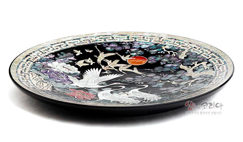 [텍스트: 샵오브코리아 대한민국의 품격까지 전합니다.] 흑색 옻칠 바탕 위 자개 상감 기법으로 송학도와 일월, 모란과 구름 문양을 섬세하게 표현한 원형 나전칠기 쟁반 모습