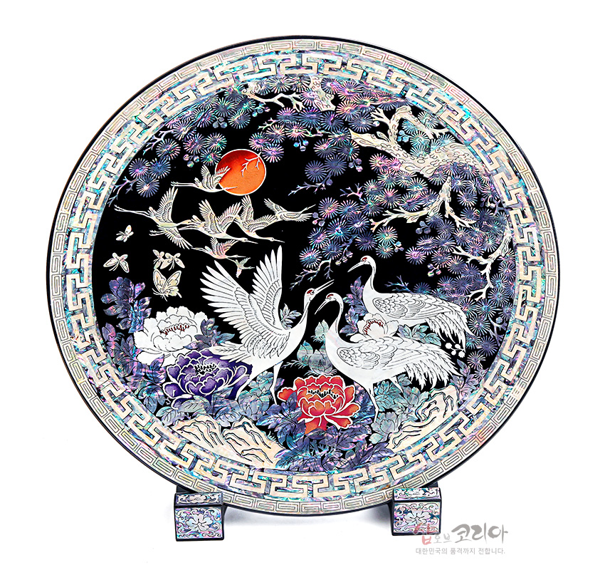 [텍스트: 코리아 대한민국의 품격까지 전합니다.] 흑색 옻칠 바탕 위 자개 상감 기법으로 송학도, 모란, 소나무와 만월 문양을 섬세하게 표현한 원형 나전칠기 쟁반 장식품