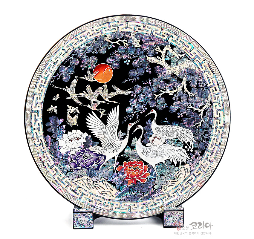[텍스트: 코리아 대한민국의 품격까지 전합니다.] 흑색 옻칠 바탕에 자개 상감 기법으로 송학도, 해바라진 붉은 해, 모란과 소나무, 구름·학 무늬를 오색으로 표현한 원형 나전칠기 쟁반 장식품