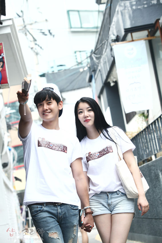 [텍스트: 샵오브코리아] 흰색 면 티셔츠에 갈색 톤 한글과 하회탈 문양 입체발포 프린트 장식, 캐주얼한 거리 배경 연출