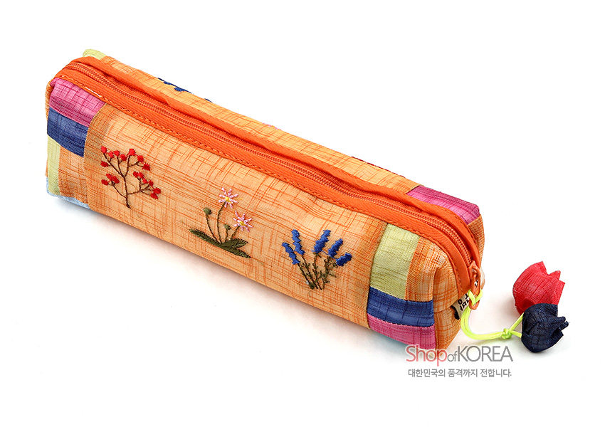 [텍스트: ShopofKOREA 대한민국의 품격까지 전합니다.] 황색 모시 천 위에 꽃자수 장식과 색동 조각천 배색, 조개 모양 전통 매듭 장식이 어우러진 필통 디자인