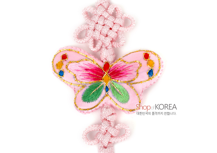 [텍스트: Shop of KOREA 대한민국의 품격까지 전합니다.] 분홍색 비단 바탕에 금사로 테두리 장식된 나비 자수 문양과 연분홍 매듭 기법이 조화를 이룬 전통 노리개 클로즈업 이미지 설명