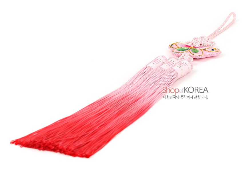 [텍스트: Shop of KOREA 대한민국의 품격까지 전합니다.] 분홍색 매듭 위 자수 나비 장식과 연분홍에서 진홍색으로 그라데이션된 실 술이 조화를 이룬 전통 노리개 이미지 설명