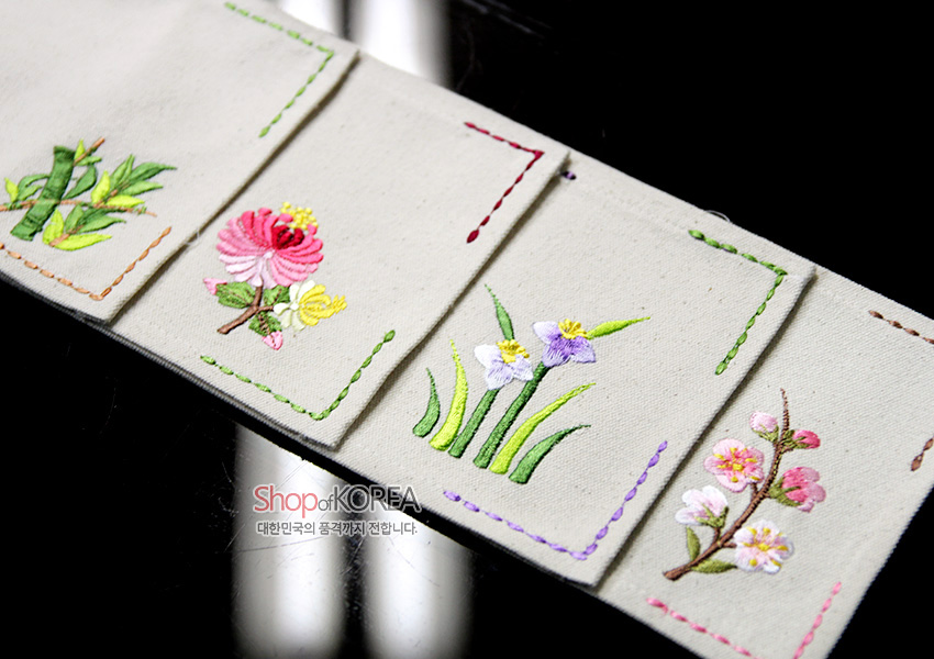 [텍스트: Shop of KOREA 대한민국의 품격까지 전합니다.] 광목 천 위에 대나무·국화·붓꽃·매화 등 꽃문양을 색실 자수로 수놓고 가장자리를 스티치 선으로 장식한 전통 자수 패브릭 식탁보 디테일 장면