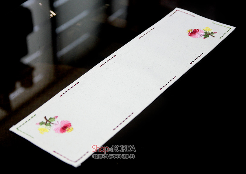 [텍스트: Shop of KOREA 대한민국의 좋은것까지만 모았습니다] 광목 재질 직사각형 식탁보, 가장자리 컬러 스티치와 양끝 분홍·노랑 국화 자수 문양 장식 이미지 설명