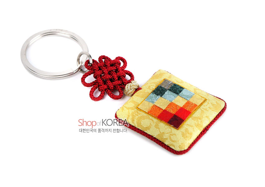 [텍스트: Shop of KOREA 대한민국의 품격까지 전합니다.] 노란 비단 바탕 사각 거울에 다채로운 조각보 패치워크 장식, 붉은 전통 매듭과 금속 키링이 연결된 열쇠고리형 공예품
