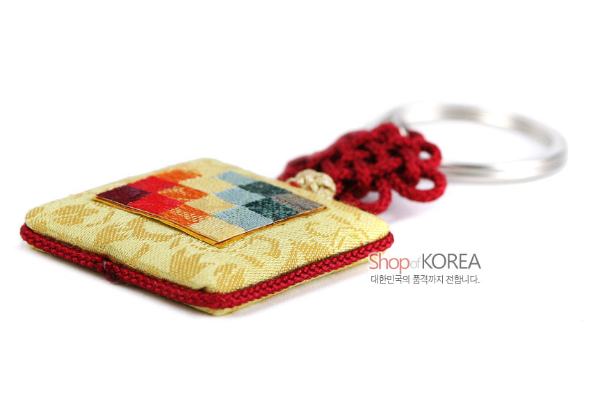 [텍스트: Shop of KOREA 대한민국의 품격까지 전합니다.] 황색 비단 바탕에 전통 문양이 직조된 사각 패드와 다색 조각보 패치워크, 붉은 전통 매듭과 금속 링이 어우러진 열쇠고리형 거울 장식
