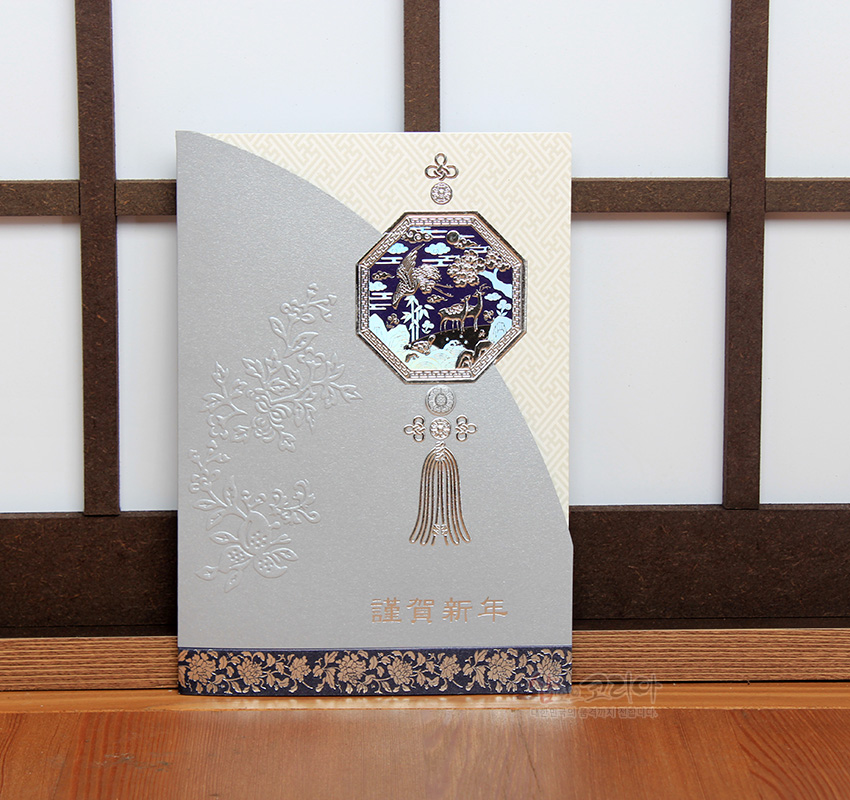 [텍스트: 謹賀新年] 은은한 은색 카드지와 아이보리 배경 위에 양각 꽃무늬, 금박 노리개 매듭, 청색·금색 산수·학·사슴 문양 장식 연하장
