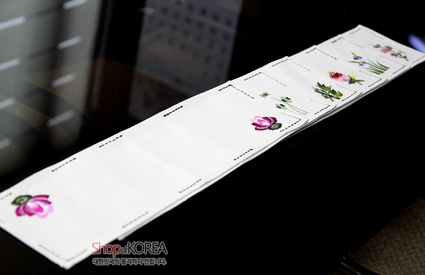 [텍스트: Shop of KOREA 대한민국의 품격까지 전합니다] 광목 재질 아이보리 긴 직사각 식탁보, 분홍 연꽃과 다양한 꽃 자수 문양, 가장자리 색실 스티치 장식 디자인