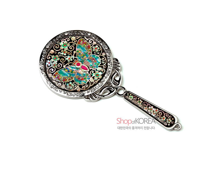 [텍스트: ShopofKOREA 대한민국의 품격까지 전합니다.] 자개 상감 기법으로 채색나비와 꽃문양을 올린 금속 테두리 손거울, 손잡이에 자개와 곡선무늬 장식 이미지 설명