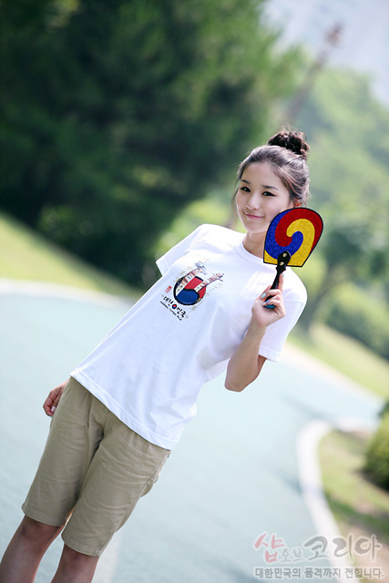 [텍스트: 샵오브코리아 대한민국의 품격까지 전합니다] 흰색 면 티셔츠 중앙 장승 형태와 태극 문양 3D 입체 프린트, 손에는 적·청·황 삼색 태극부채 든 야외 연출 이미지