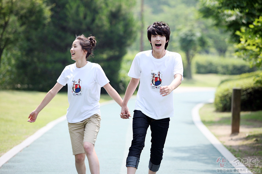 [텍스트: 샵오브코리아 대한민국의 흥을 세계까지 전합니다] 흰색 면 티셔츠 중앙에 장승 형상과 붉은·푸른 태극 문양을 입체감 있게 컬러 인쇄한 그래픽 디자인 모습