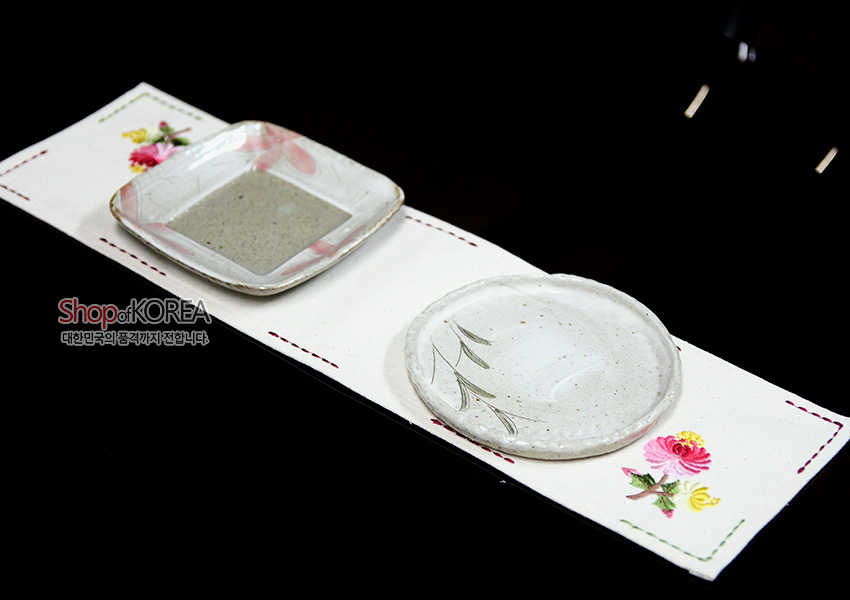 [텍스트: Shop of KOREA 대한민국의 품격까지 전합니다] 광목 재질 긴 러너형 식탁보 위 자수 사군자 꽃문양과 컬러 스티치 가장자리 장식, 도자기 접시 연출 이미지