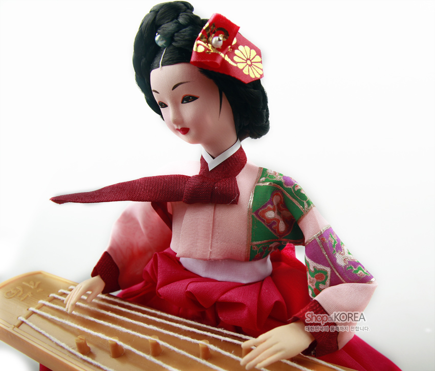 [텍스트: Shop KOREA 대한민국의 품격까지 전합니다.] 분홍 비단 치마와 패치워크 문양 저고리를 입고, 나무 결이 살아있는 가야금을 연주하는 전통 한복 인형 장식