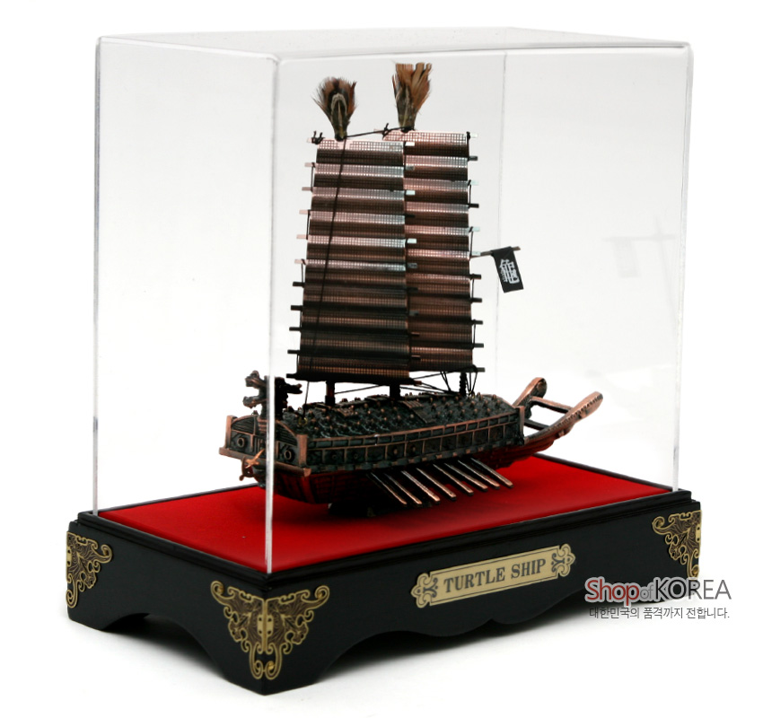 [텍스트: TURTLE SHIP, Shop of KOREA] 금속 질감의 거북선 모형을 투명 아크릴 케이스에 배치하고, 붉은색 바닥과 흑색 목재 받침, 금색 장식 문양으로 구성한 장식품 이미지