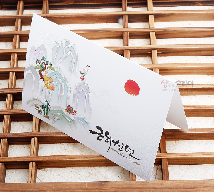 [텍스트: 근하신년 Season's Greetings] 수묵화풍 산수와 소나무, 사슴, 학, 대나무 길상문양을 파스텔 톤으로 인쇄한 백색 종이 재질 전통 연하장 디자인