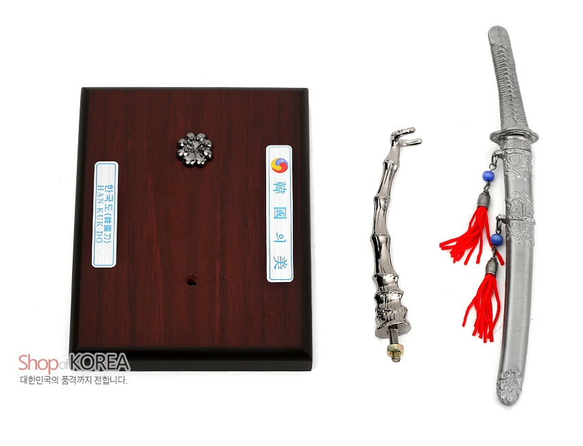 [텍스트: 한국도(韓國刀) HAN-KUKDO, 韓國의 美] 금속 재질의 한국도 모형과 음각 문양 장식 검집, 붉은 술과 청색 구슬 매듭, 대나무 형상의 금속 스탠드와 나무 받침대 구성 이미지