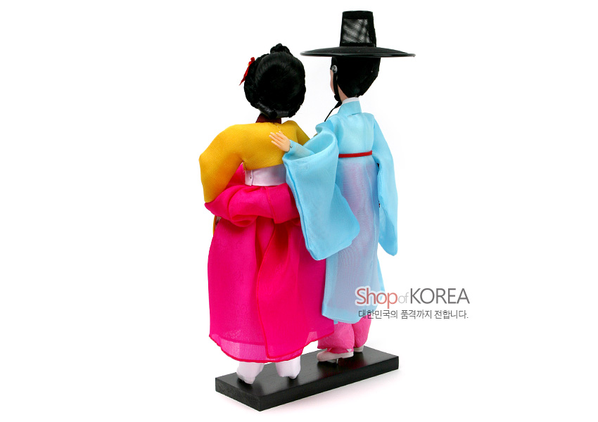 [텍스트: Shop of KOREA 대한민국의 품격까지 전합니다.] 비단 느낌의 선명한 분홍·노랑 치마저고리와 하늘색 도포, 흑립을 쓴 선비와 기녀 한복 인형 한 쌍 뒷모습 연출 이미지