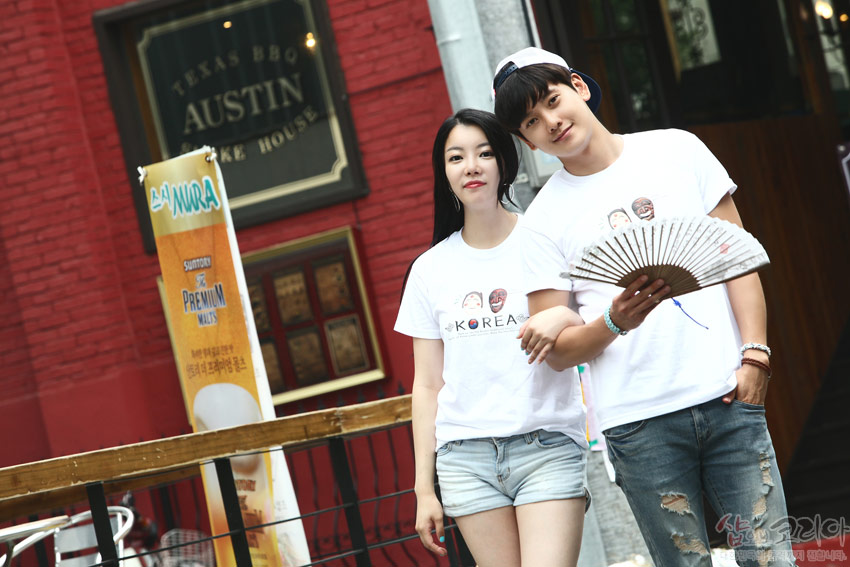 [텍스트: KOREA] 흰색 면 티셔츠 전면에 하회탈 캐릭터와 영문 레터링이 3D 입체발포 기법으로 인쇄된 캐주얼 커플 착용 이미지