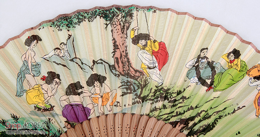 [텍스트: ShopKOREA 대한민국전통기념품샵] 옅은 연두 바탕 종이 부채면에 신윤복 단오도 풍속 인물과 수목, 그네 장면을 다색 채색으로 표현한 합죽선 형 접이식 부채 이미지