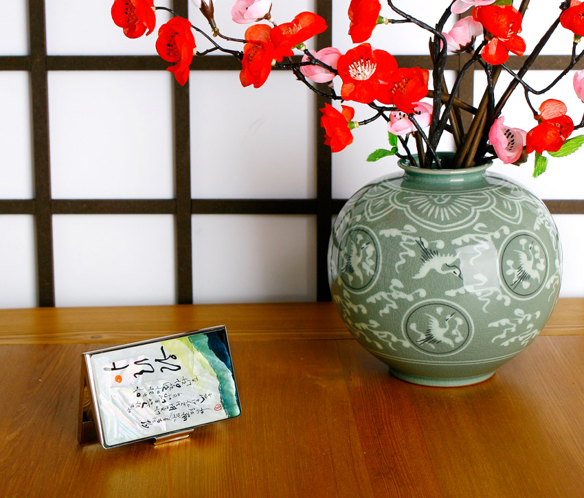 [텍스트: 아리랑] 금속 재질 위 녹청·청록 그라데이션 수묵담채 배경과 아리랑 가사 서예가 어우러진 직사각형 명함집 이미지 설명