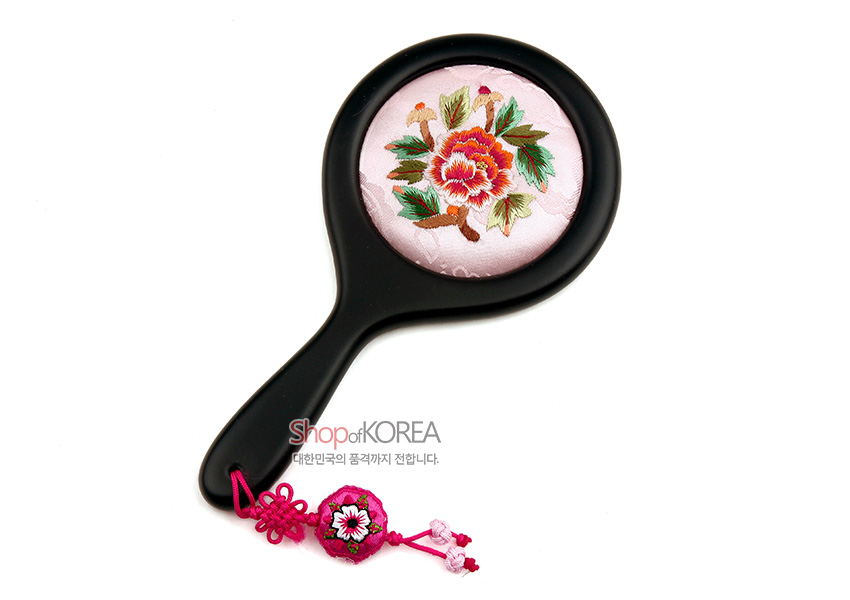 [텍스트: ShopofKOREA 대한민국의 품격까지 전합니다.] 연분홍 비단 바탕 위 모란 자수 문양 원형 손거울과 검정 손잡이, 분홍 매듭과 꽃 자수 장식 매듭고리 구성