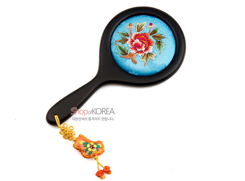[텍스트: Shop of KOREA 대한민국의 품격까지 전합니다.] 하늘색 비단 바탕에 화려한 꽃 자수 문양이 놓인 원형 손거울과 노란 매듭 장식이 달린 복주머니형 술 장식 구성
