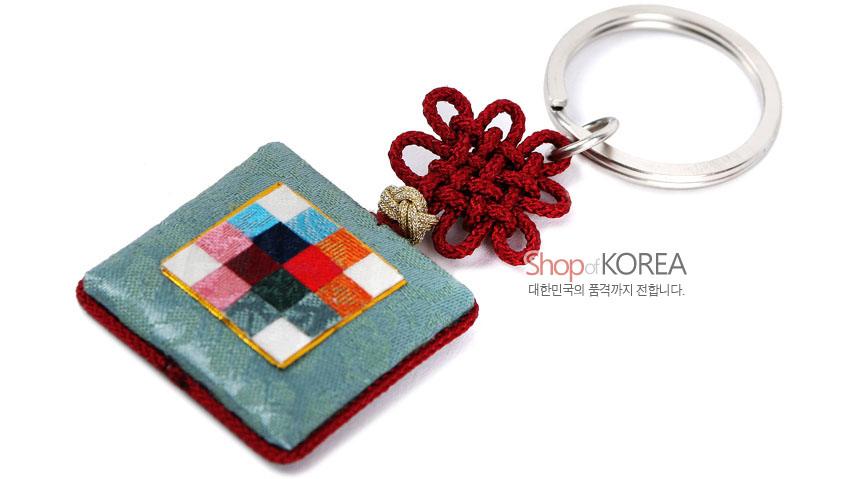 [텍스트: Shop of KOREA / 대한민국의 품격까지 전합니다.] 옥색 비단 방석형 패드 위 다색 조각보 패턴 장식, 붉은 전통 매듭과 금속 링이 조화된 거울 열쇠고리 디자인