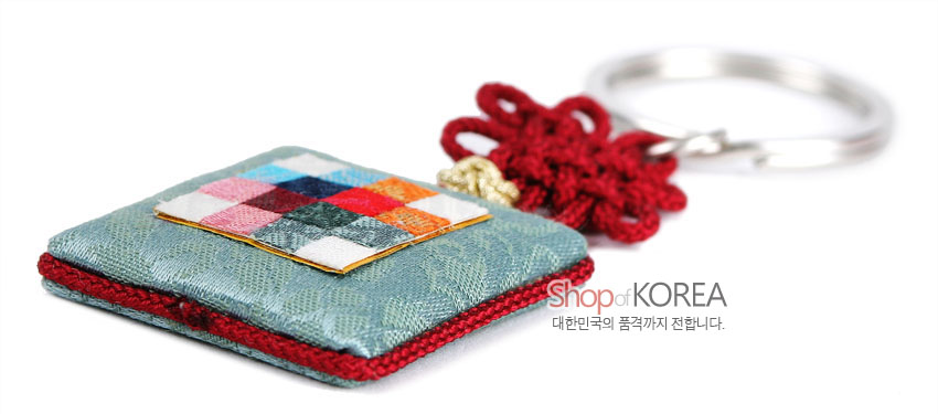 [텍스트: Shop of KOREA 대한민국의 품격까지 전합니다.] 옥색 비단 방석형 패드 위 다색 조각보 패턴 장식과 붉은 전통 매듭, 금속 고리가 어우러진 열쇠고리 이미지 설명