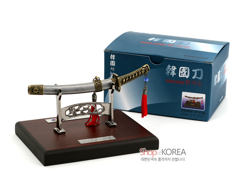 [텍스트: 韓國刀, Shop of KOREA, STAND STYLE Made in KOREA] 금속 재질 한국도 모형에 전통 문양 조각과 흑금색 칼손잡이, 붉은 술 장식, 목재 받침대가 어우러진 스탠드형 장식품