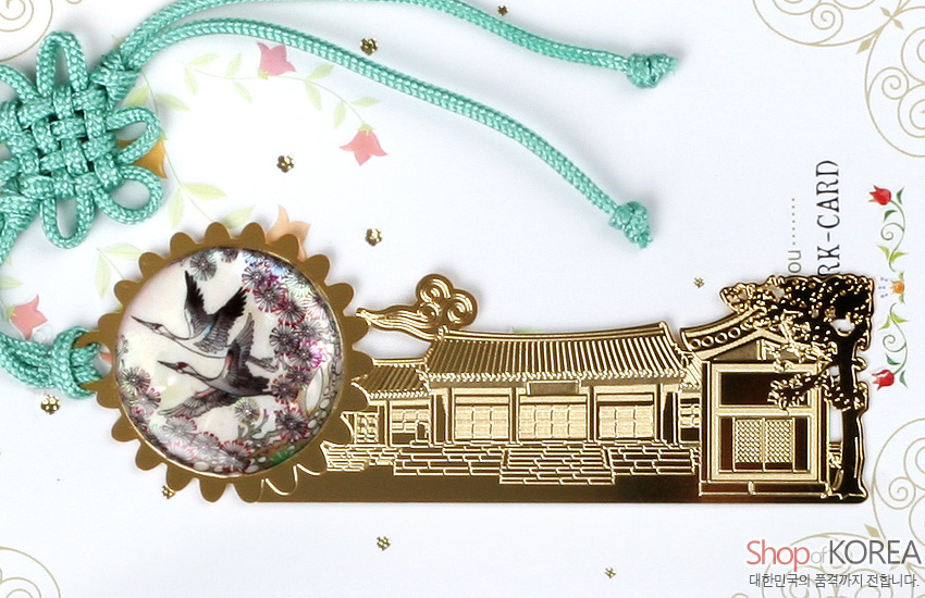 [텍스트: Shop of KOREA, 대한민국의 품격까지 전합니다.] 금속 재질 기와집 실루엣에 쌍학과 매화 자개 원형 장식, 청록색 전통 매듭 끈 포인트의 금장 책갈피 이미지 설명