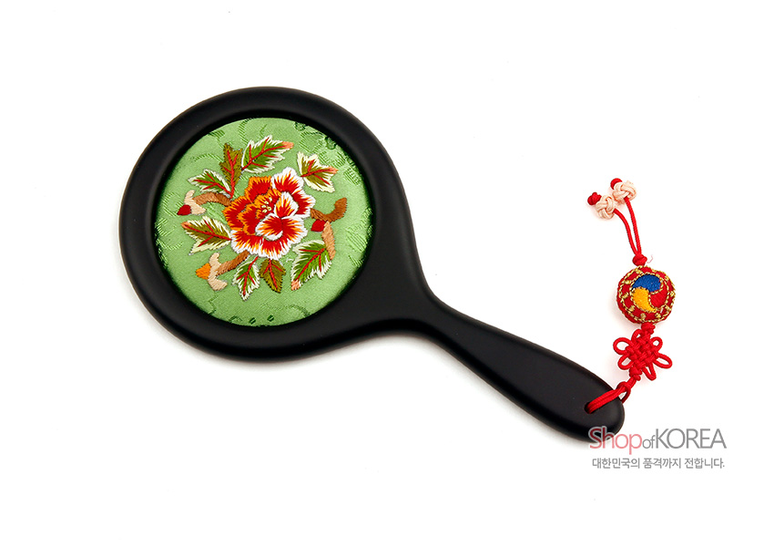 [텍스트: ShopofKOREA 대한민국의 품격까지 전합니다] 연두 비단 바탕 위 붉은 모란 자수 장식 원형 거울과 흑색 손잡이, 붉은 전통 매듭과 조롱박 매듭 장식 손거울 모습