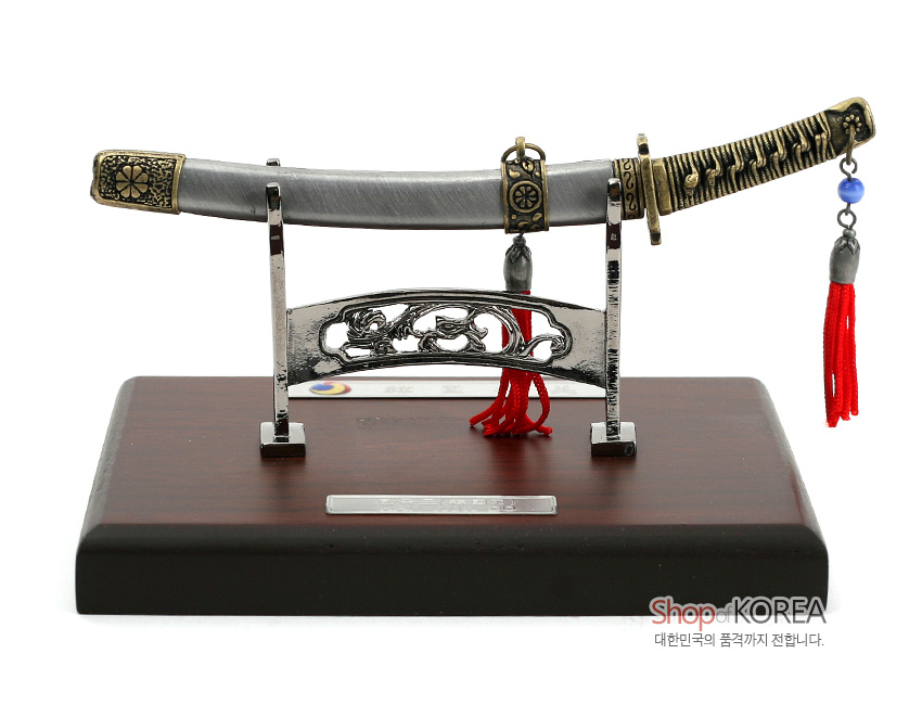 [텍스트: ShopKOREA 대한민국의 품격까지 전합니다.] 금속 재질 칼집과 칼자루에 전통 문양 양각 장식, 붉은 술과 청색 구슬 매듭 장식, 목재 받침대와 금속 스탠드가 조화를 이룬 한국도 모형 장식품