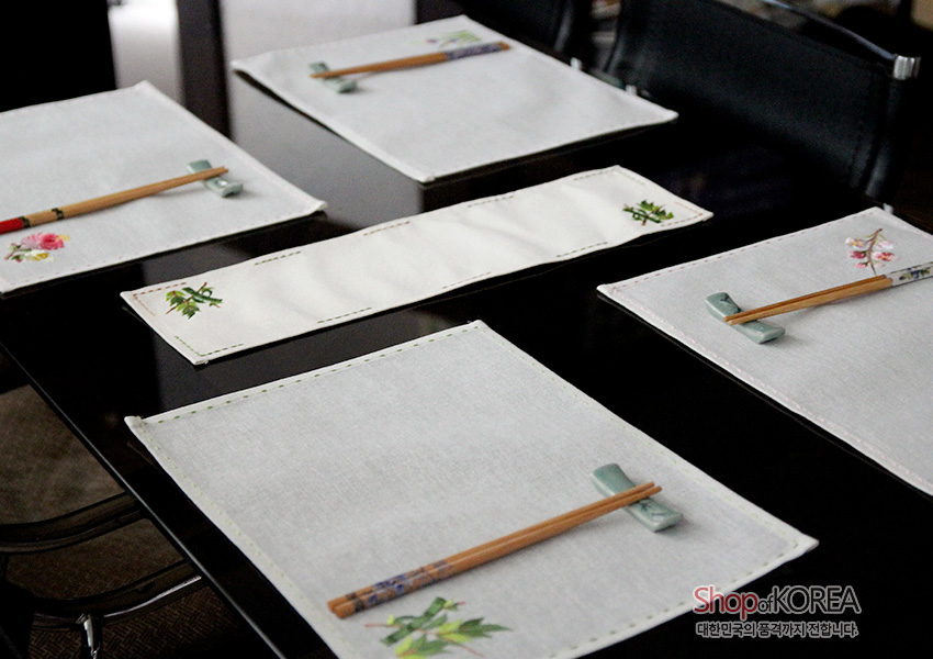[텍스트: Shop of KOREA 대한민국의 품격까지 전합니다] 광목 재질 아이보리 식탁보 세트 위에 연꽃·매화 등 식물 자수와 가장자리 스티치가 더해진 단정한 상차림 연출 이미지