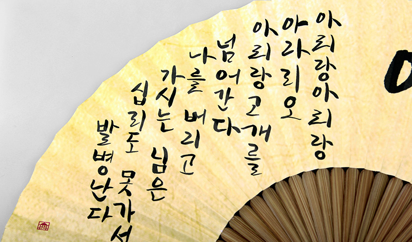 [텍스트: 아리랑 아리랑 아라리요 아리랑 고개를 넘어간다 나를 버리고 가시는 님은 십리도 못가서 발병난다] 연한 황토색 한지 바탕에 먹색 한글 아리랑 가사 서예와 대나무 살이 어우러진 전통 부채 클로즈업 이미지