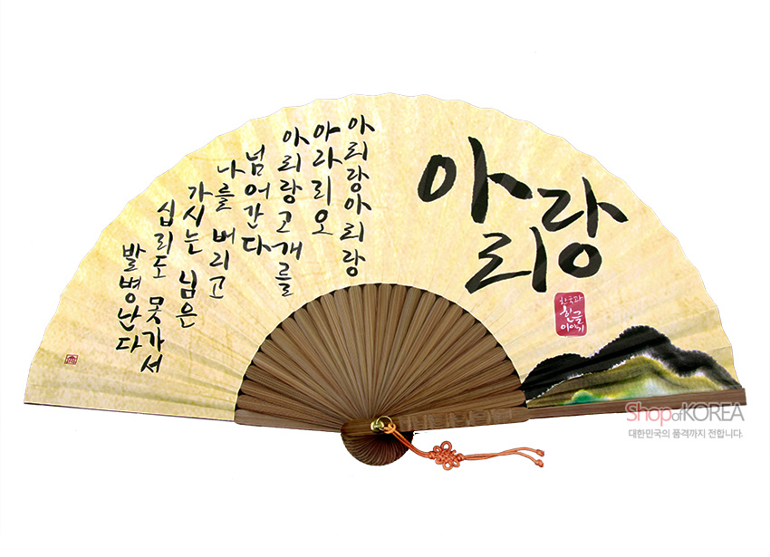 [텍스트: 아리랑 아리랑 아라리요 아리랑 고개를 넘어간다 나를 버리고 가시는 님은 십리도 못가서 발병난다 / 아리랑 / 샵오브코리아] 연한 황토색 한지 바탕에 먹색 한글 서예와 산수화가 어우러진 합죽선, 갈색 대나무살과 주황 전통 매듭 장식 배치