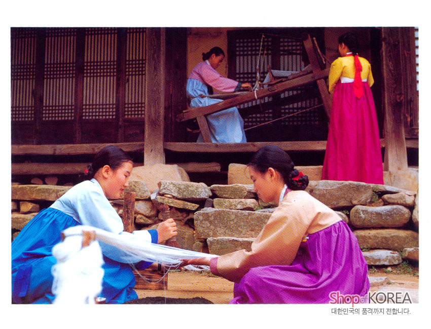 [텍스트: Shop of KOREA / 대한민국의 품격까지 전합니다.] 전통 한옥 마당에서 푸른색·분홍색·보라색 비단 한복 차림 여인들이 베를 짜고 실을 잣는 풍경엽서 이미지