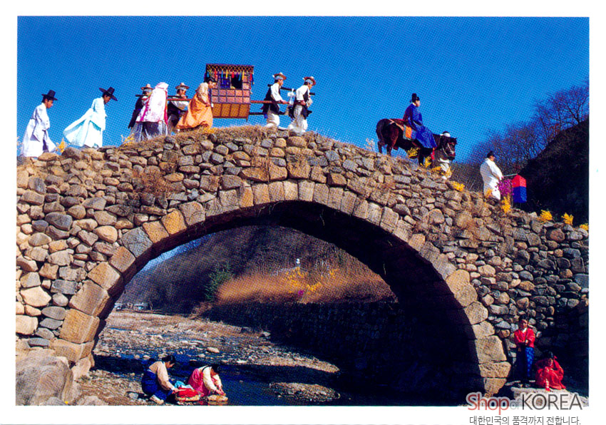 [텍스트: Shop of KOREA 대한민국의 품격까지 전합니다.] 돌다리 위 행렬과 가마, 말을 탄 인물들의 다채로운 비단 한복, 아래 냇가의 전통 일상 풍경을 담은 사진 엽서 이미지 설명