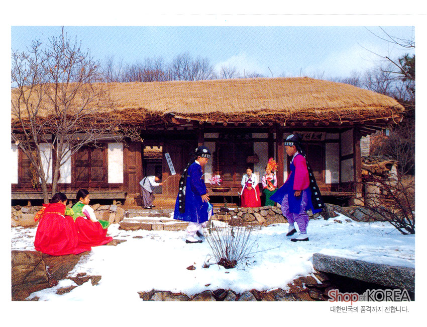 [텍스트: Shop OF KOREA / 대한민국의 품격까지 전합니다.] 겨울 눈 덮인 초가집 마당에서 비단 한복과 색동 옷을 차려입은 인물들이 전통 놀이를 즐기는 풍경 엽서 이미지 묘사