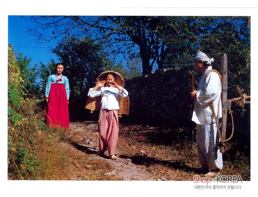 [텍스트: Shop KOREA / 대한민국의 품격까지 전합니다.] 파란 하늘 아래 돌담길에서 비단 한복과 전통 생활복을 입은 인물들이 서 있는 한국 농촌 풍경 엽서 이미지 묘사