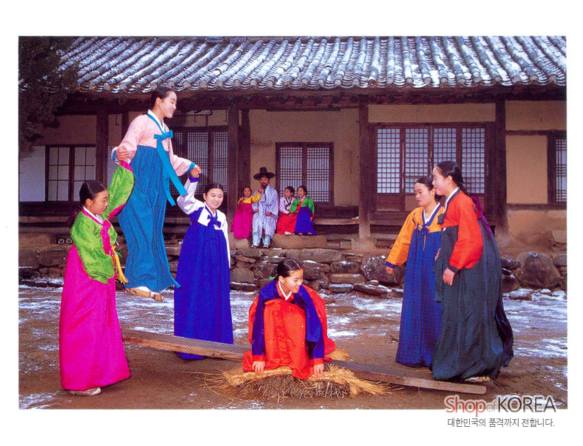 [텍스트: Shop KOREA / 대한민국의 품격까지 전합니다.] 전통 한옥 마당에서 색동·단색 비단 한복을 입은 인물들이 널뛰기와 윷놀이를 즐기는 겨울 풍경 엽서 이미지